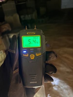 A moisture meter reading 5.4% is being used to measure moisture content on what appears to be a basement or crawlspace