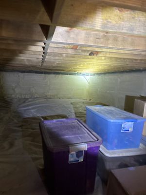 A crawlspace under construction showing plastic storage containers staged on the floor, exposed wooden joists above,