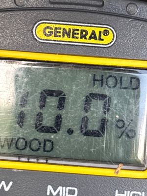 A moisture meter displays a wood moisture reading of 0.0% while positioned on what appears to be a foundation wall or