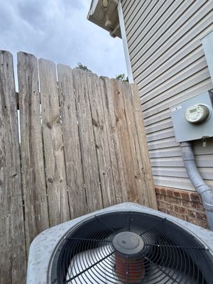 Exterior view of an HVAC condenser unit positioned against a wooden fence next to a residential building with metal