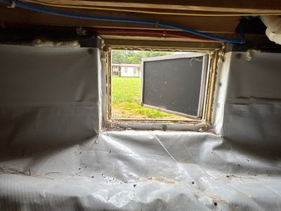 A crawlspace window opening with white vapor barrier material being installed on the interior walls and floor during