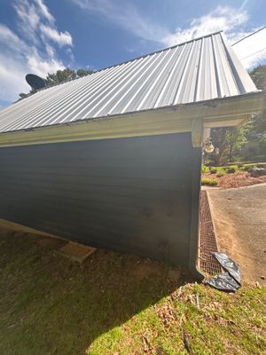 A large black plastic drainage basin or catch basin is being installed in the yard beneath a metal-roofed structure,
