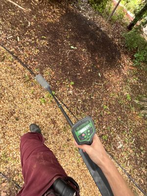 A technician holds a moisture meter over wet soil during an exterior yard drainage assessment or installation, with