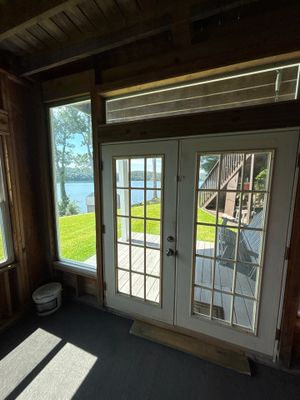 Interior view of a sunroom or enclosed porch with French doors and windows overlooking a lawn and water, showing no