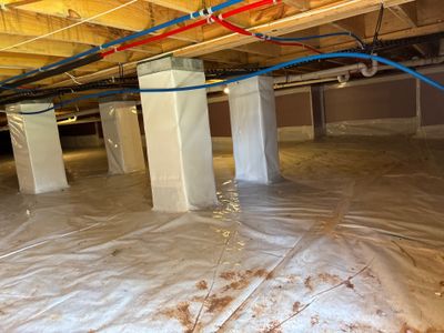 Crawlspace encapsulation installation in progress with vapor barrier sheeting, support posts, and blue drainage piping