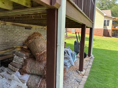 Foundation repair staging area under a deck showing stacked drainage materials, gravel base preparation, and yard