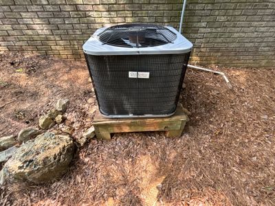 An HVAC condenser unit is positioned on a wooden pallet next to a foundation with visible yard drainage installation or