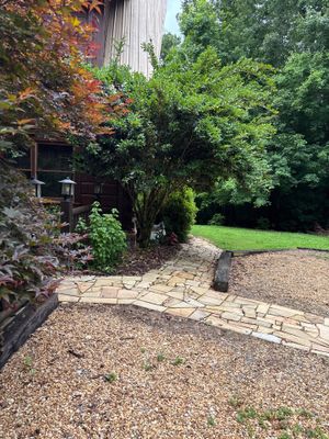 A residential property exterior showing a brick foundation, gravel yard area, stone patio, and manicured landscaping