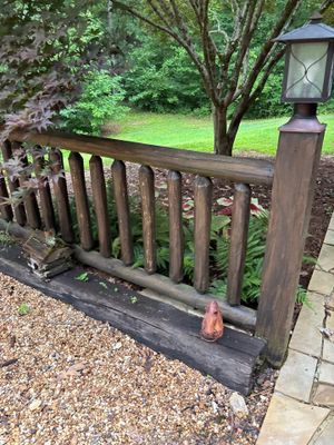 A residential deck or porch with wooden railings and posts overlooking a landscaped yard with mature trees, showing no