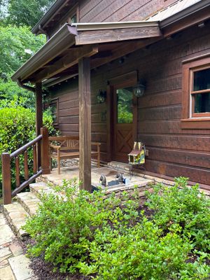 A residential wooden cabin exterior with a covered porch and surrounding landscaping, showing no visible foundation