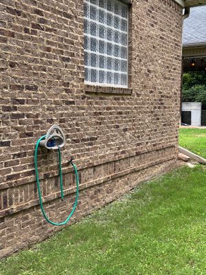 Exterior brick foundation wall with a green yard-drainage hose connected to a wall-mounted spigot, showing the pre-work
