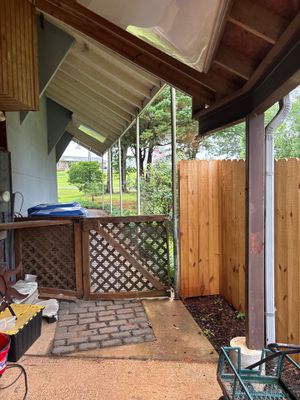 A covered porch or patio area with brick/stone flooring, wooden lattice panels, and yard furniture visible, showing the