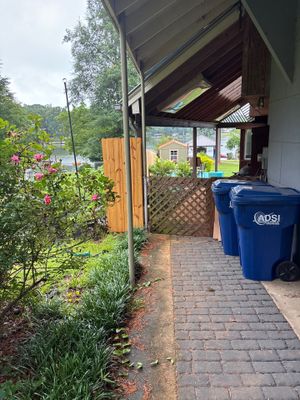 Exterior view of a residential home's side entrance with a covered walkway, brick pavers, landscaping, and trash bins,