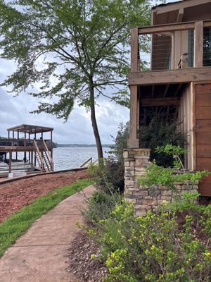 A waterfront residential property with a brick foundation, wooden deck structure, and mature trees, showing no visible