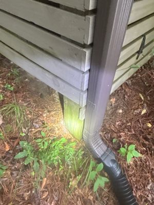 A black drainage pipe with elbow fitting is installed at the foundation base, directing water away from the house into