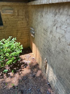 A narrow exterior foundation space between two concrete walls with vegetation and mulch visible, showing the pre-work