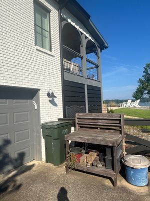 A waterfront residential property showing the exterior foundation and yard area with an old wooden bench, green trash