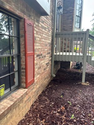 Exterior foundation area with brick walls, red louvered vent, gray deck/railing, and dark mulch ground surface showing