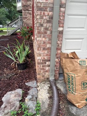 Exterior foundation area showing installed drainage downspout extension with flexible corrugated pipe running along the