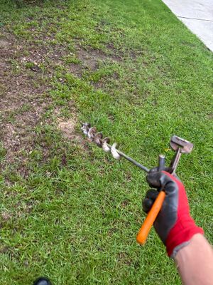 A worker holding a trenching tool is actively digging or preparing yard drainage in a lawn area, with visible soil