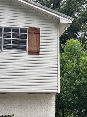 The image shows the exterior of a white-sided residential building with a wooden shutter and window, adjacent to dense