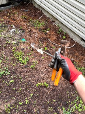 A person wearing work gloves is installing or working on exterior yard drainage in a landscaped bed next to a metal