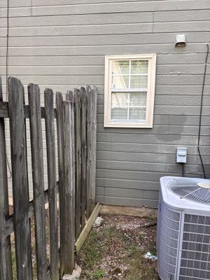 Exterior view of a house foundation showing a weathered wooden fence, gray fiber cement siding, a white-trimmed window,