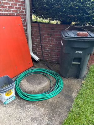 Exterior job site showing staged drainage materials including a green garden hose coiled on concrete, a black plastic