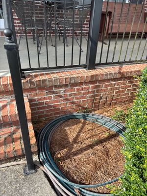 Exterior yard drainage setup showing a coiled black drainage pipe staged next to a brick foundation wall with metal