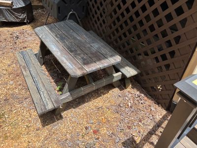 A weathered picnic table sits on gravel next to a brown lattice fence, showing an outdoor recreational area with no