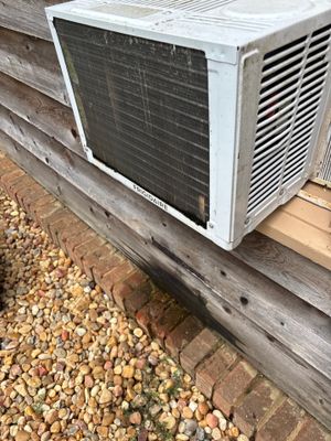 An exterior window air conditioning unit mounted on a foundation with gravel landscaping visible below, showing the