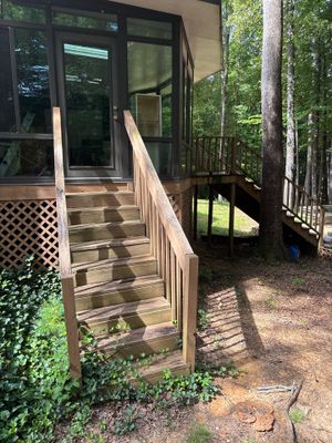 Exterior view of a residential deck and entrance with wooden stairs and railings, surrounded by landscaping and trees,