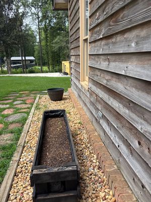 Exterior foundation drainage trench with black plastic drain channel installed along the base of a wooden house