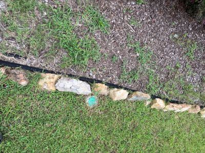Exterior yard drainage installation in progress with a black drain line laid across a grass lawn with stone/aggregate