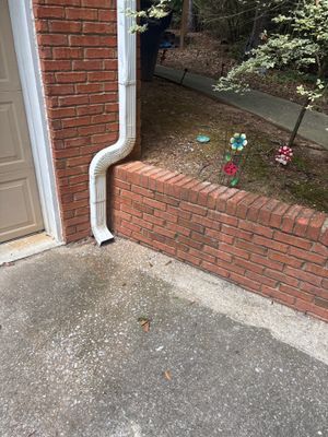Exterior foundation view showing a brick wall with white downspout and landscaped flower beds with visible drainage