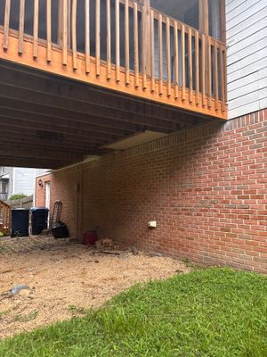 Exterior view of a brick foundation with a deck above and exposed ground/lawn area below showing the pre-work condition