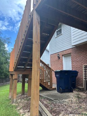 A brick residential home with a wooden deck structure and a black trash bin visible, showing the exterior foundation