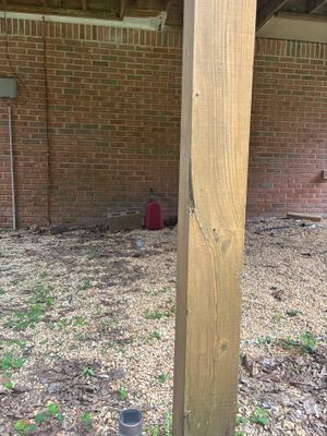 A wooden support post is visible in an excavated area behind a brick foundation with a black drainage basin or