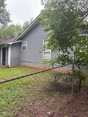 A gray single-story house with overgrown vegetation and a chain-link fence in the yard, showing no visible foundation