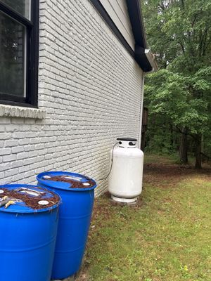 Exterior foundation area showing two blue drums and a white tank positioned near the house foundation with forested