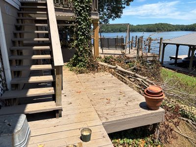 A waterfront residential property showing a wooden deck and stairs with landscaping, dock infrastructure, and water