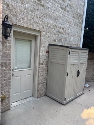 A residential exterior showing a brick wall with a white entry door and a gray plastic storage shed, with no visible