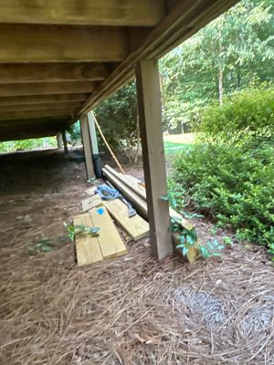 Exterior crawl space or foundation area under a wooden structure showing staged drainage materials and equipment