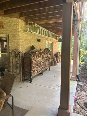 A covered porch or breezeway with brick walls, wooden support columns, and stacked firewood, showing no visible