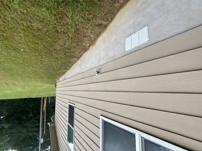 Exterior view of a residential home with tan/beige horizontal siding, showing the foundation area and lawn, with no
