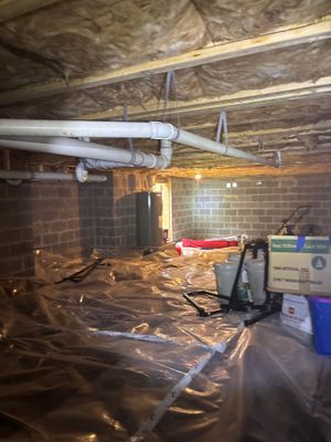 A crawl space with black vapor barrier membrane being installed across the floor and interior drainage piping visible