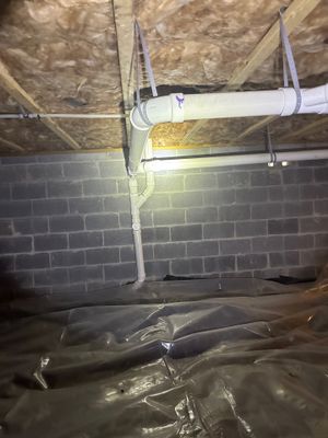 A crawlspace encapsulation project in progress showing a white PVC drainage pipe suspended from the foundation wall