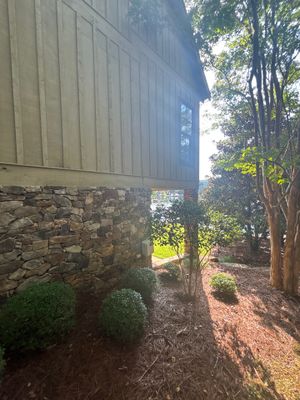 Exterior view of a house foundation with stone base and siding, flanked by landscaping and trees, showing no visible