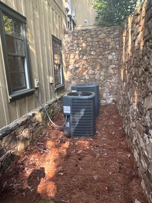 An exterior HVAC unit is positioned in a narrow side yard alley with fresh mulch/wood chips spread on the ground,