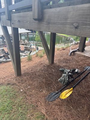 Exterior under-deck area with yard drainage equipment (black drain box and yellow pipe) staged on mulch ground during